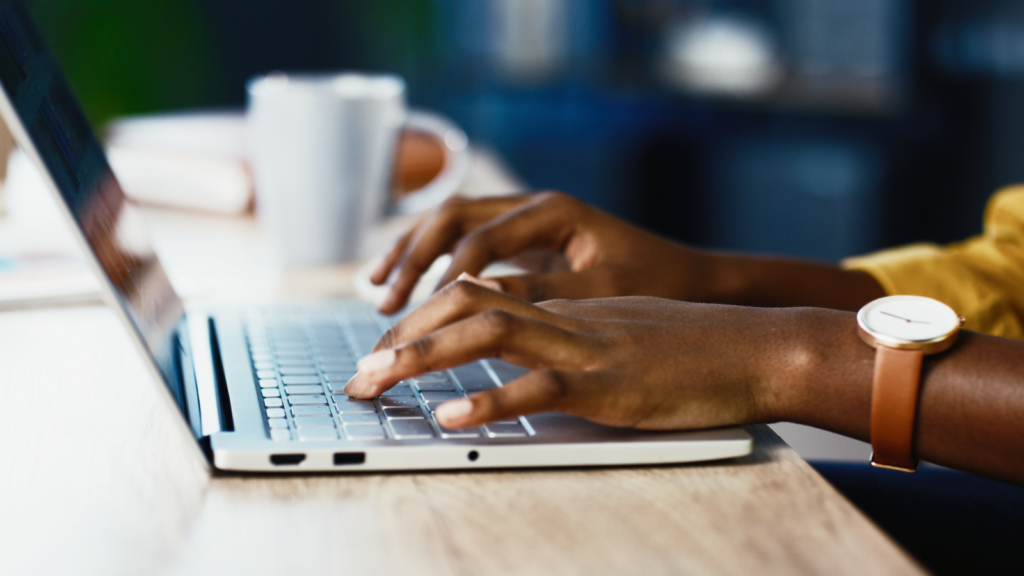 Hands typing on a laptop keyboard, with a focus on a wristwatch. A blurred mug and notebook in the background suggest a workspace setting.