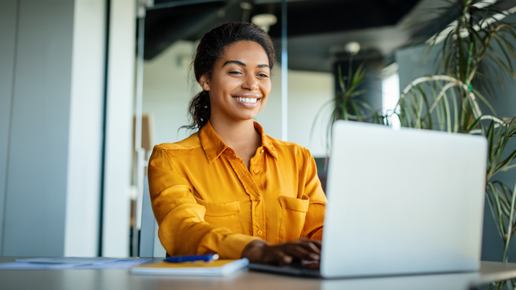 Smiling woman in a bright orange shirt using a laptop in a modern office with plants. Her expression conveys confidence and positivity.