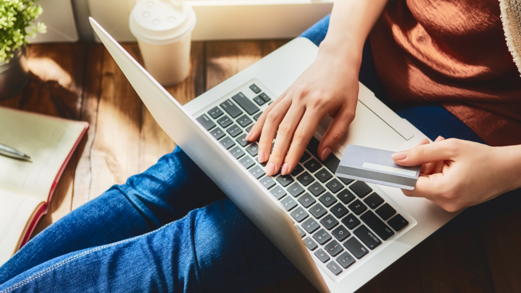 A person in jeans sitting on a wooden floor uses a laptop while holding a credit card, suggesting online shopping.