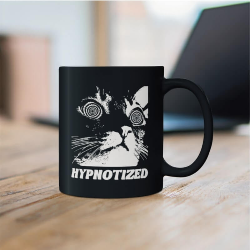 Print On Demand With Shopify Mugs A black mug with an image of a cat with swirls for eyes and the text “Hypnotized.”