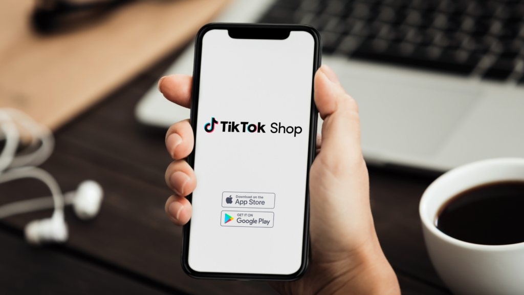 A hand holds a smartphone displaying the TikTok Shop logo, with icons for downloading on the App Store and Google Play. 