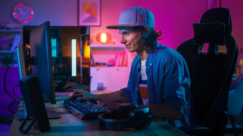 A young man sitting by his desk with a full gaming and streaming setup, in a room with soft neon lighting.
