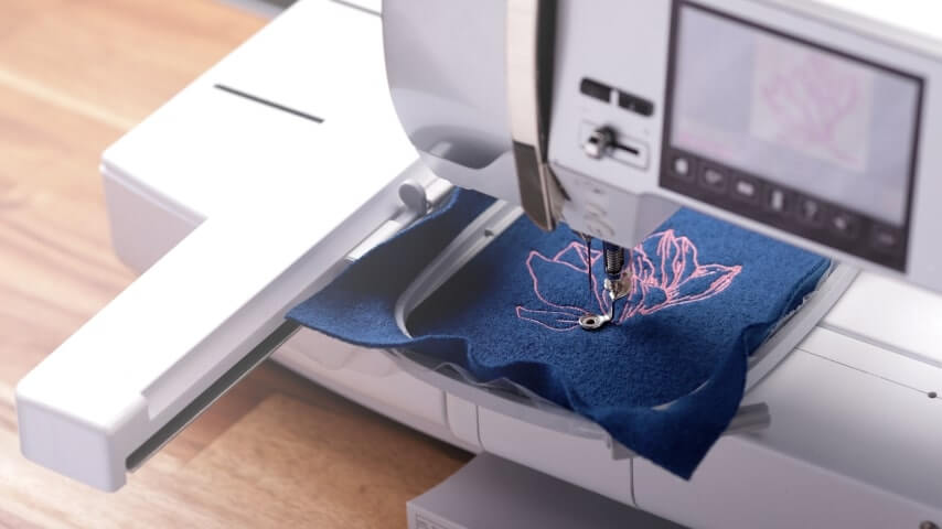 A sample piece of blue fabric being embroidered with a pink flower design.