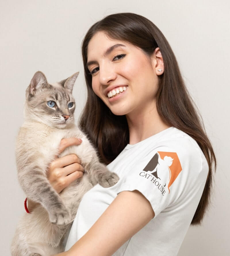 A woman holding a cat, wearing a white t-shirt with a sleeve print of a logo that says, “Cat House.”