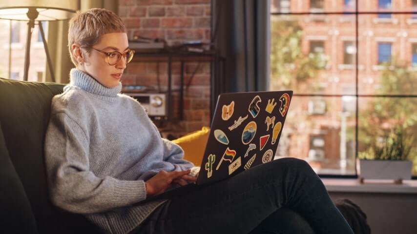 A person wearing glasses and a cozy sweater is comfortably working on a sticker-adorned laptop in a well-lit room with a city view.