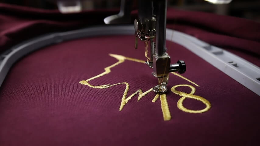 A maroon garment being embroidered with a golden logo design.