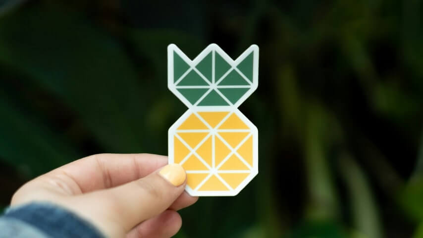 A hand holding a geometric pineapple-shaped sticker with a blurred green foliage background.