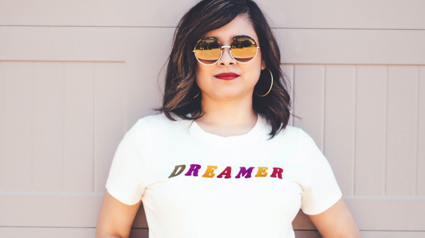 A woman in sunglasses wearing a white print-on-demand shirt with the word “Dreamer” printed on it in colorful letters.
