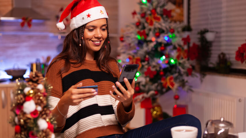 A young brunette wearing a Santa cap in a room with Christmas decorations smiles while scrolling through a smartphone with one hand and holding a credit card in the other.