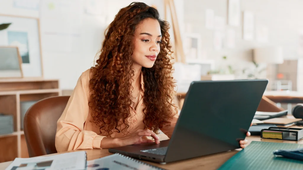 A woman with curly hair sits at a desk, focused on her laptop while working.