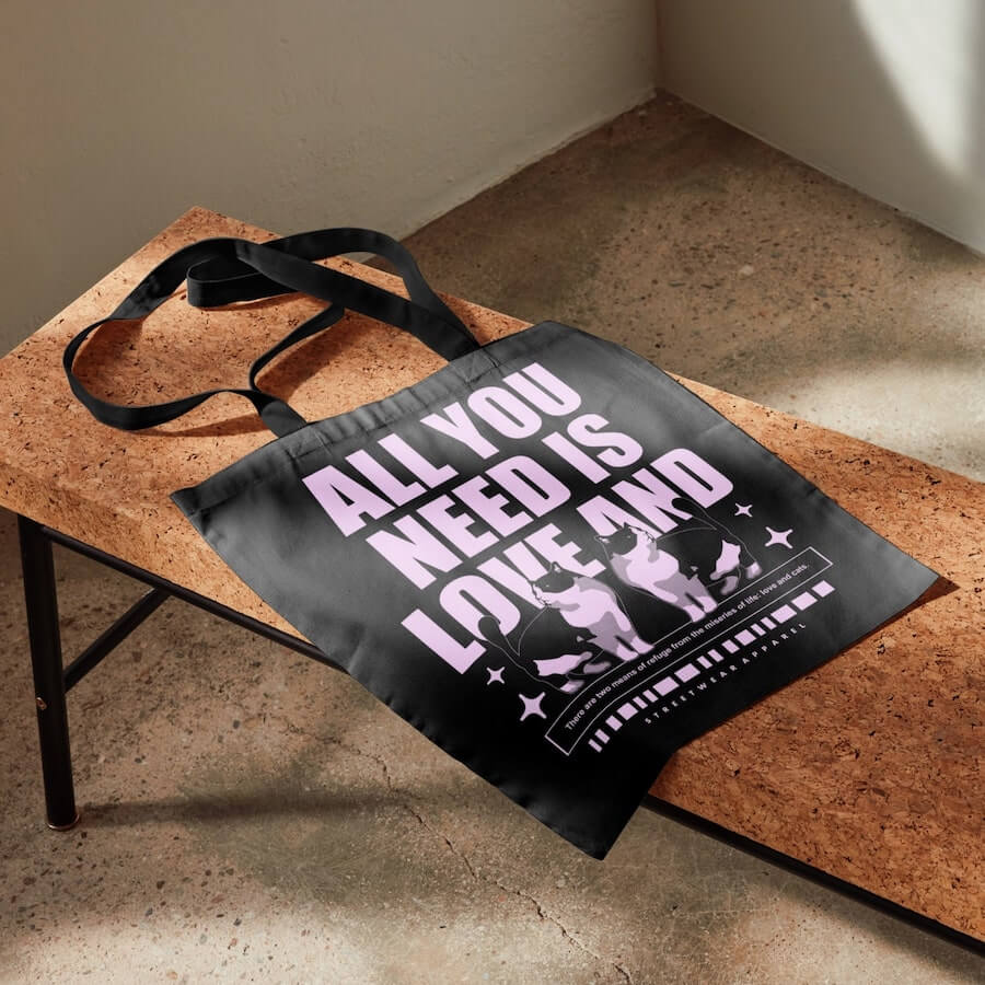 A black tote bag with the text “All you need is love and” above an image of two cats.