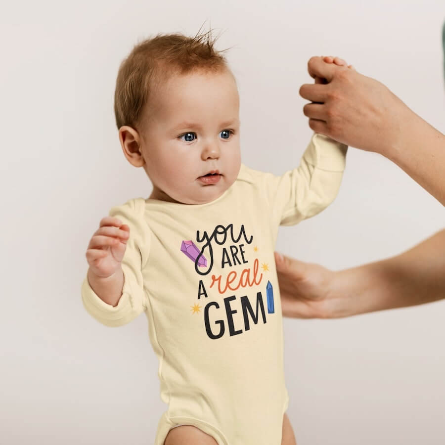 A baby wearing a yellow long-sleeve onesie with the text saying: “You are a real gem,” surrounded by a design of colorful crystals.