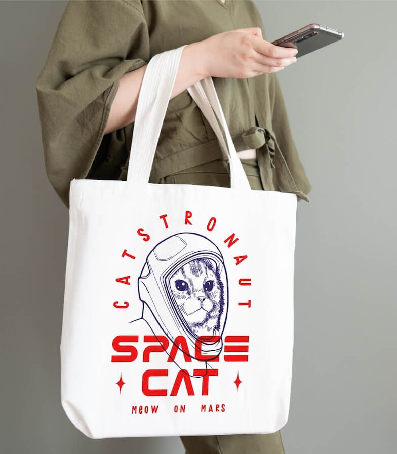 A woman holding a white tote bag with a design of a cat in a spacesuit and the text “Catstronaut. Space Cat. Meow on Mars.”