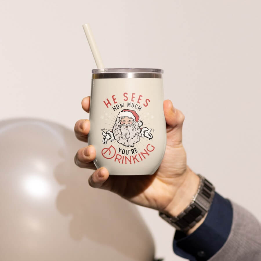 A beige tumbler with an illustration of Santa Claus and the text: “He Sees How Much You’re Drinking.”