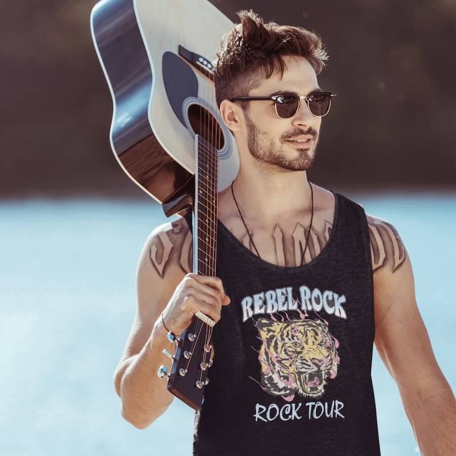 A man in sunglasses, holding a guitar, and wearing a black tank top that says “Rebel Rock. Rock Tour.”