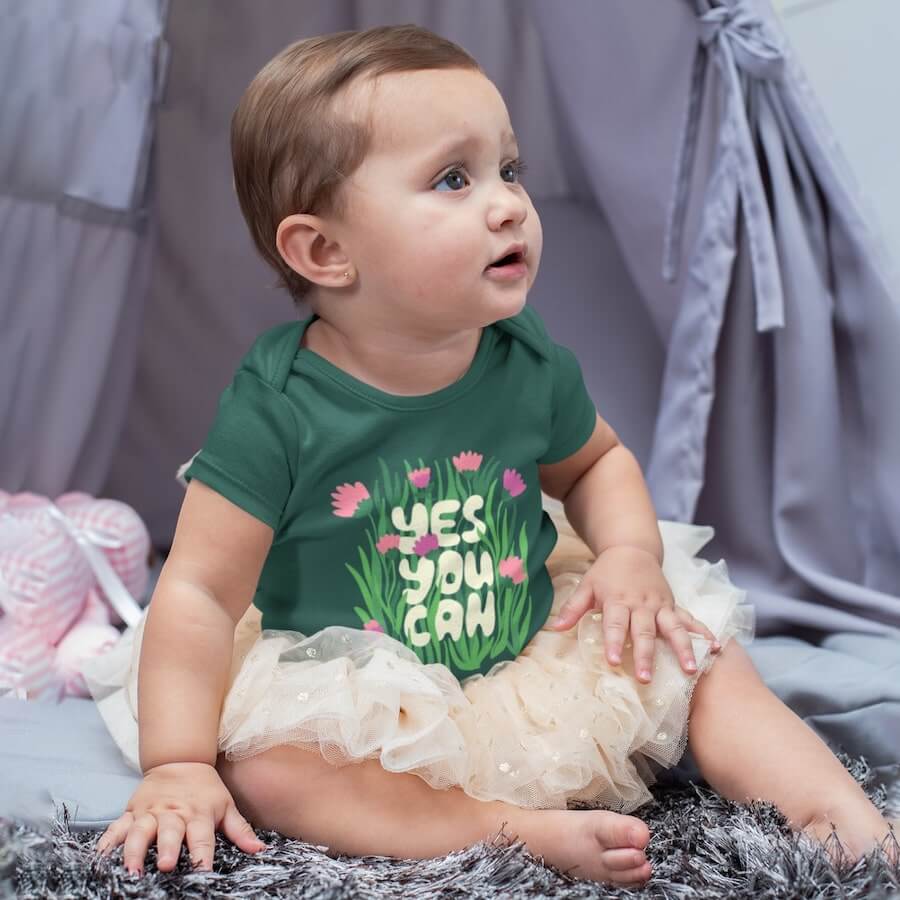 A baby wearing a green onesie with a design of flowers and the text “Yes, you can.”