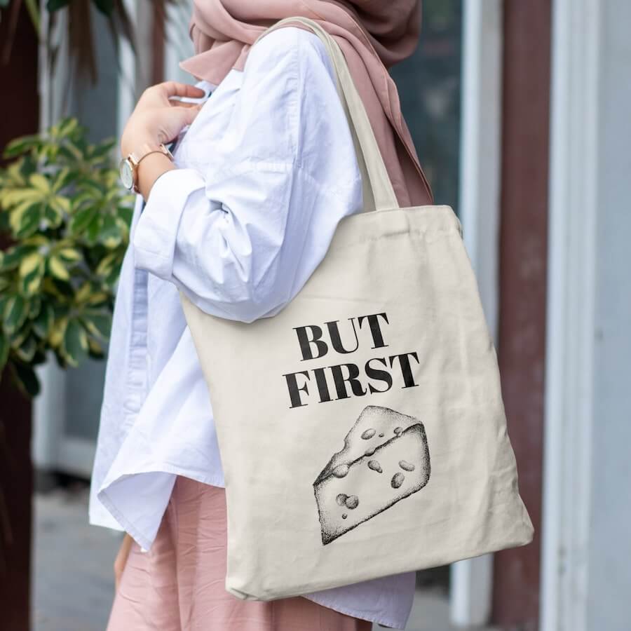 A woman with a canvas tote bag with a design of a piece of cheese and the text “But first” above it.