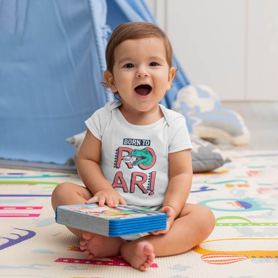 A baby boy wearing a white short-sleeve onesie with a design of a dinosaur and the text: “Born to roar.”