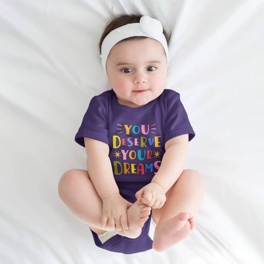 A baby girl wearing a purple onesie with a text design in colorful letters saying, “You deserve your dreams.”