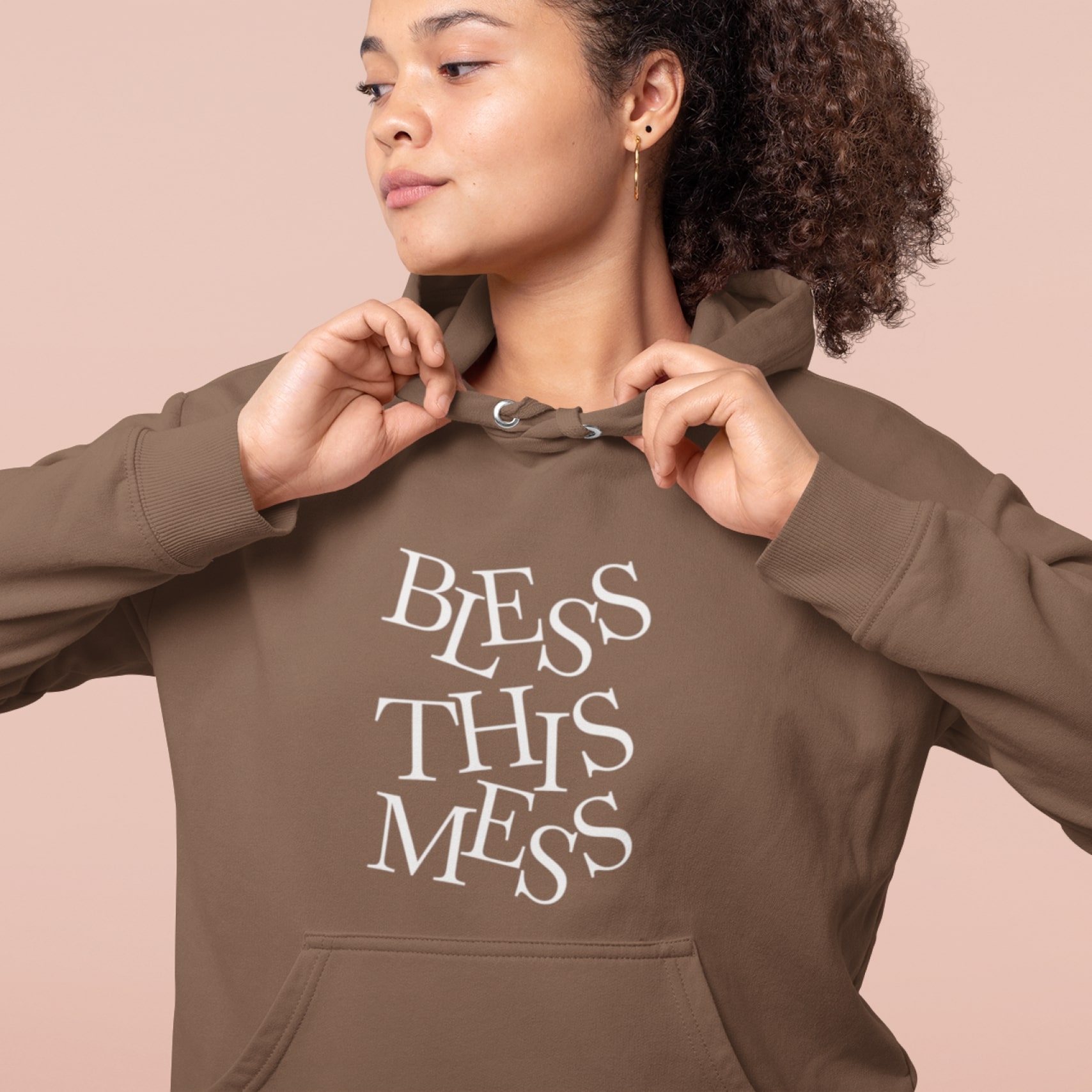 A woman wearing a custom hoodie in brown with “Bless This Mess” text on.