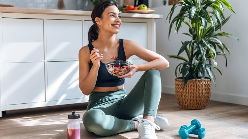 A woman wearing athletic apparel and eating a fruit salad on the floor.