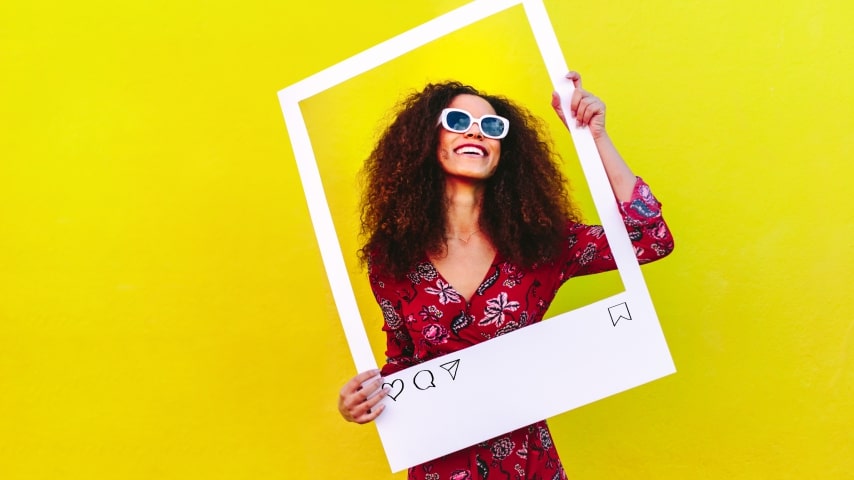A woman posing in front of a yellow background and holding up a large frame that looks like an Instagram photo border.