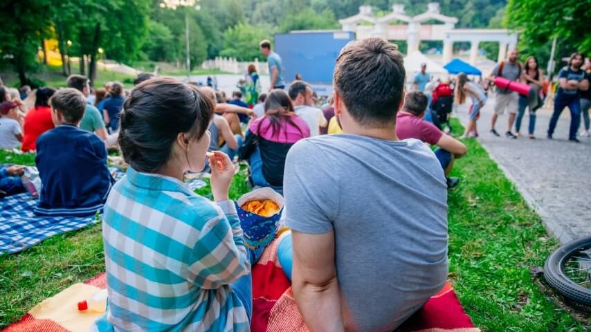 A group of people sitting on the lawn in a park, attending an outdoor movie night.