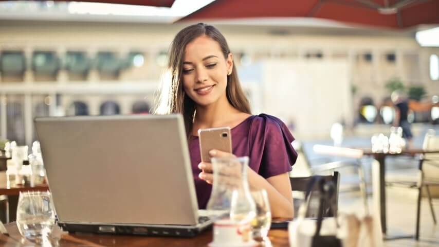 A woman holding a mobile phone while working on a laptop in a cafe or restaurant.