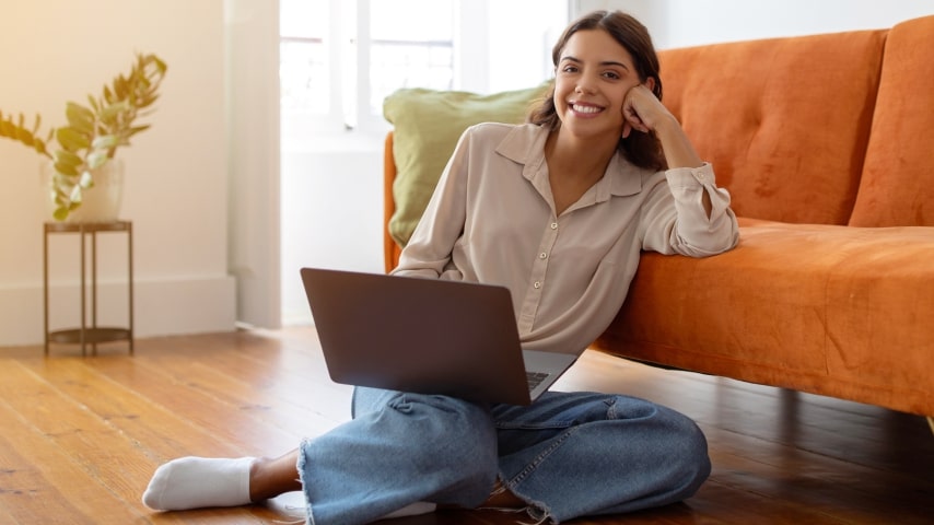 A smiling woman sits on the floor next to an orange couch with her laptop.