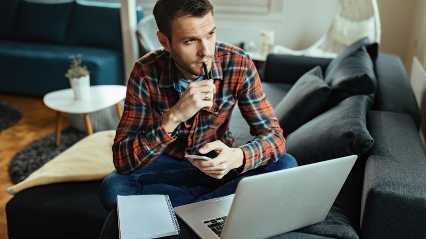 A man sitting by a laptop thinking while holding a mobile phone in his hand.