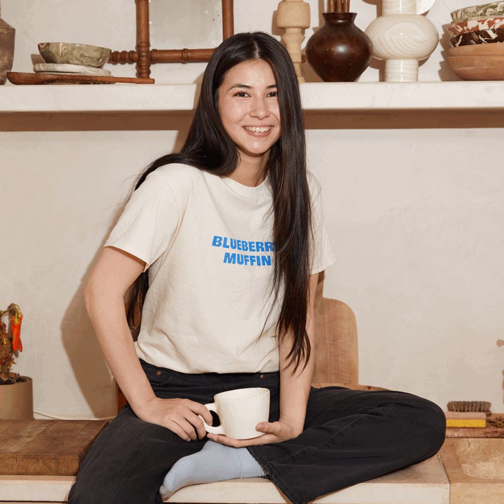 How to start a t shirt business with Print on Demand A smiling young woman wearing a beige cotton t-shirt with a blue “Blueberry muffin” text print on the front.