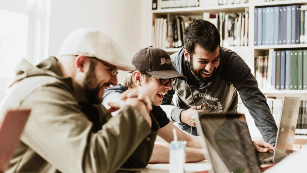 Three men are focused on their laptops while seated at a table in a quiet library setting.