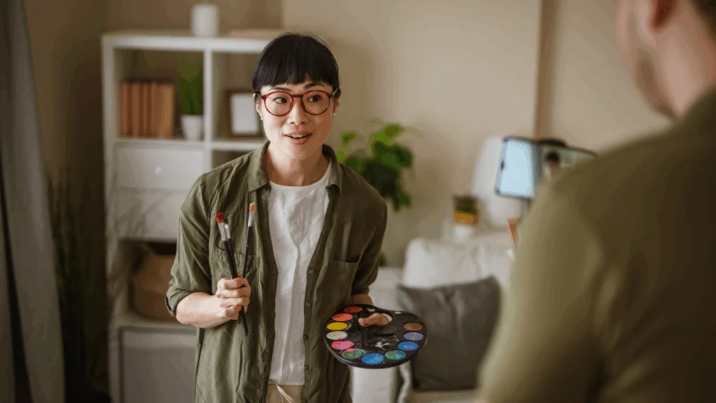 A woman artist is being recorded on video while holding paint brushes and paint.