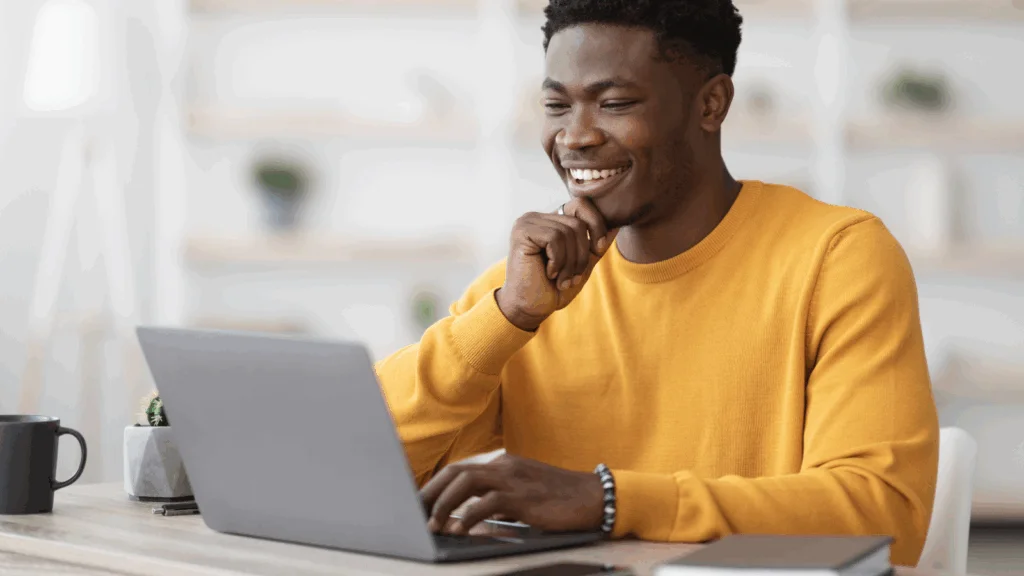 A smiling young man sits at a table, working on his laptop with a relaxed and focused expression.