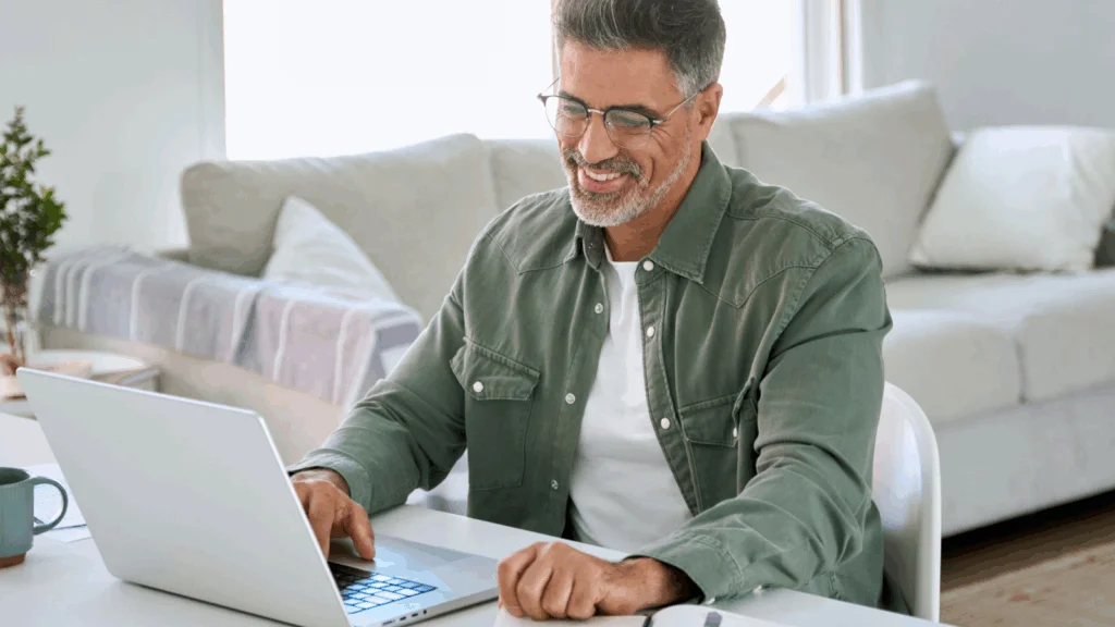 A smiling man in glasses is sitting at a desk and working on a laptop.