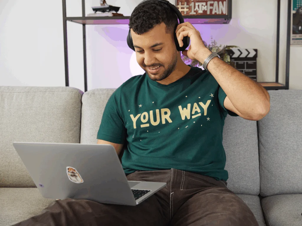 A young man in a green “Your way” t-shirt is sitting on a couch and using a laptop to find passive income ideas.