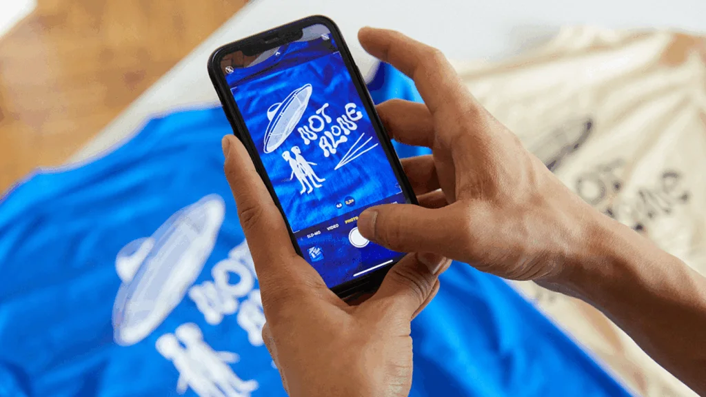 A person holding a smartphone displaying a shirt design on its screen.