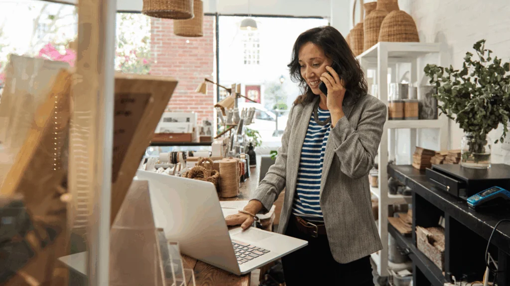A woman stands in front of a laptop, engaged in a phone conversation.