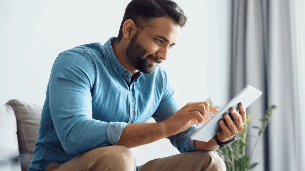 A man in a blue jeans shirt is sitting and using a tablet.