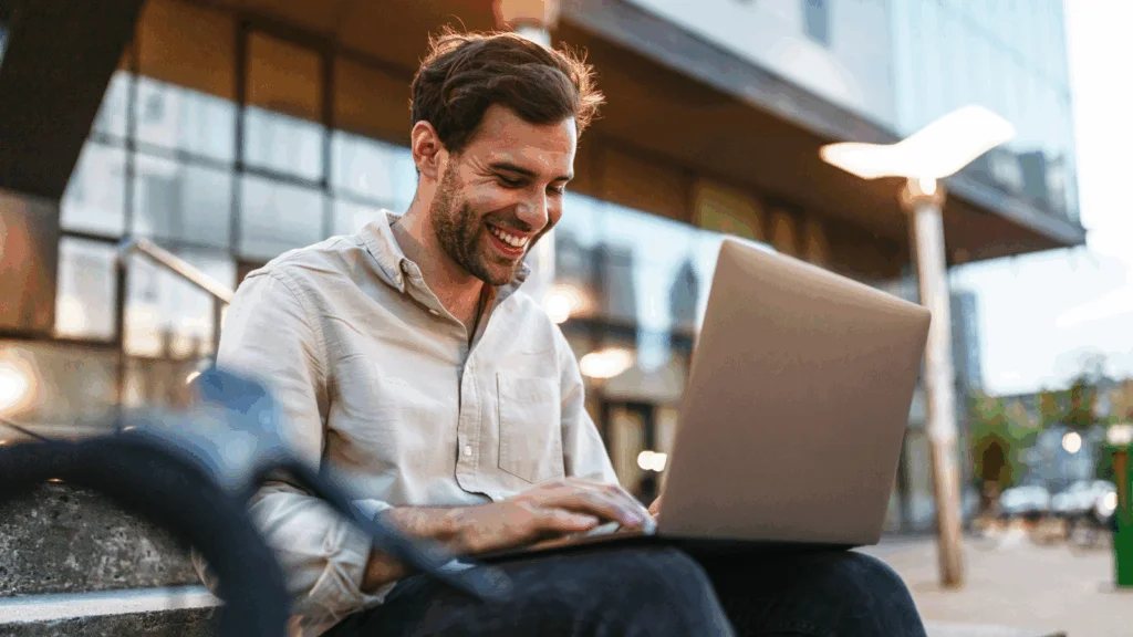 A smiling man sitting on a bench outdoors with a laptop on his lap, choosing best print-on-demand websites for artists.