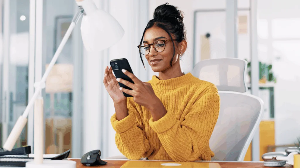 A woman in glasses and a yellow sweater is sitting and looking at a mobile phone.