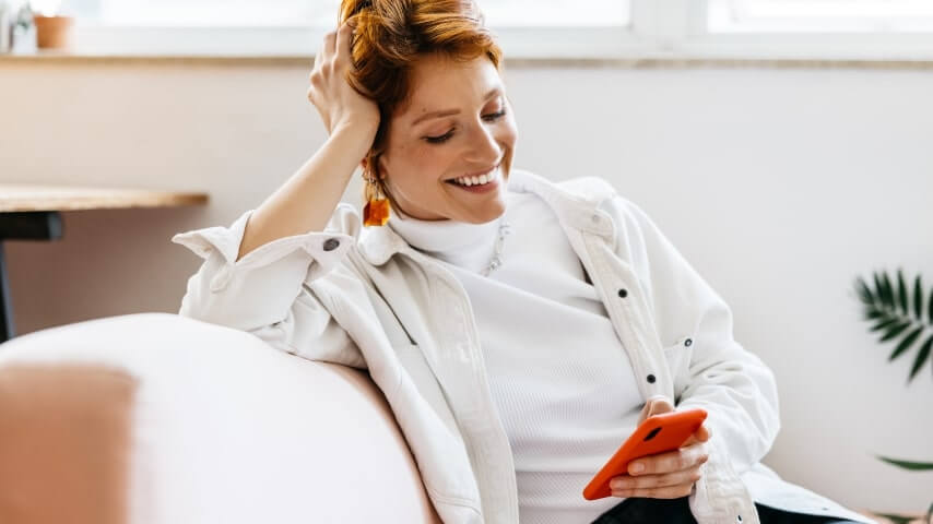 Offer great customer service A woman sitting on a couch, smiling while looking at a mobile phone in her hand.