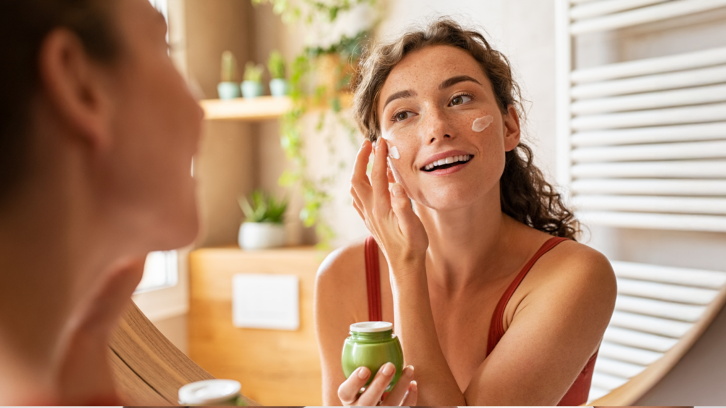 A woman applying face cream in a bright bathroom. She smiles at her reflection in the mirror. Plants are visible in the background.