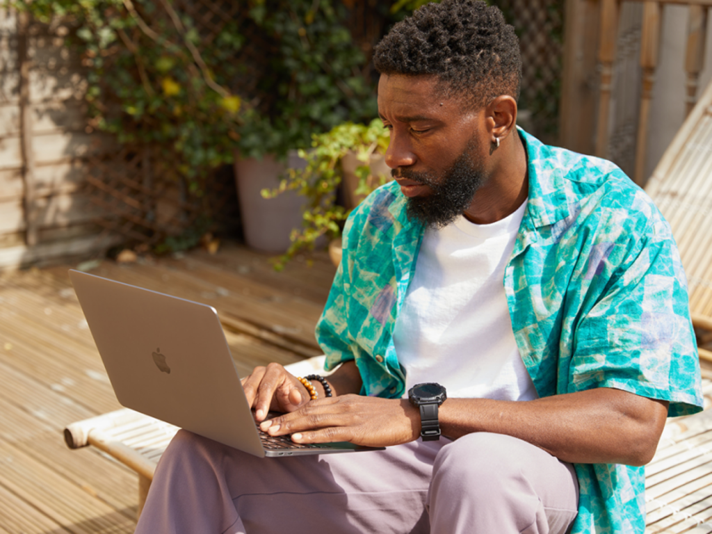 A man sitting outdoors on a wooden deck, using a laptop to search for Super Bowl marketing ideas. He wears a colorful shirt.