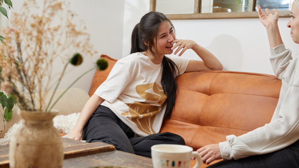 A woman in a white t-shirt laughs while sitting on a brown couch, engaging in a lively conversation with another woman.