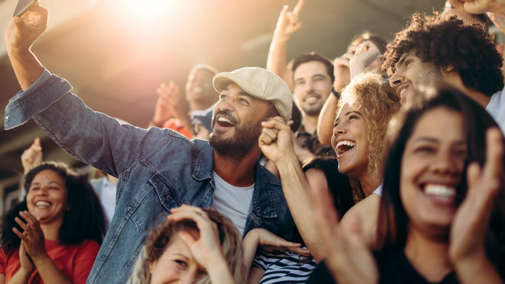 A diverse group of excited fans in a stadium, smiling and cheering. They huddle for a selfie.