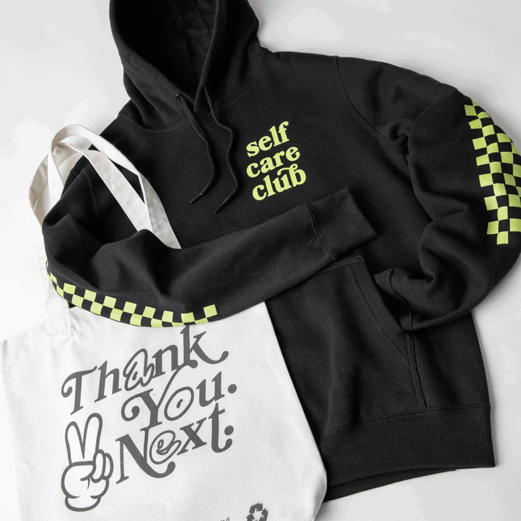 Black hoodie with "self care club" text and green checkered sleeves next to a white tote bag featuring "Thank You, Next" text.
