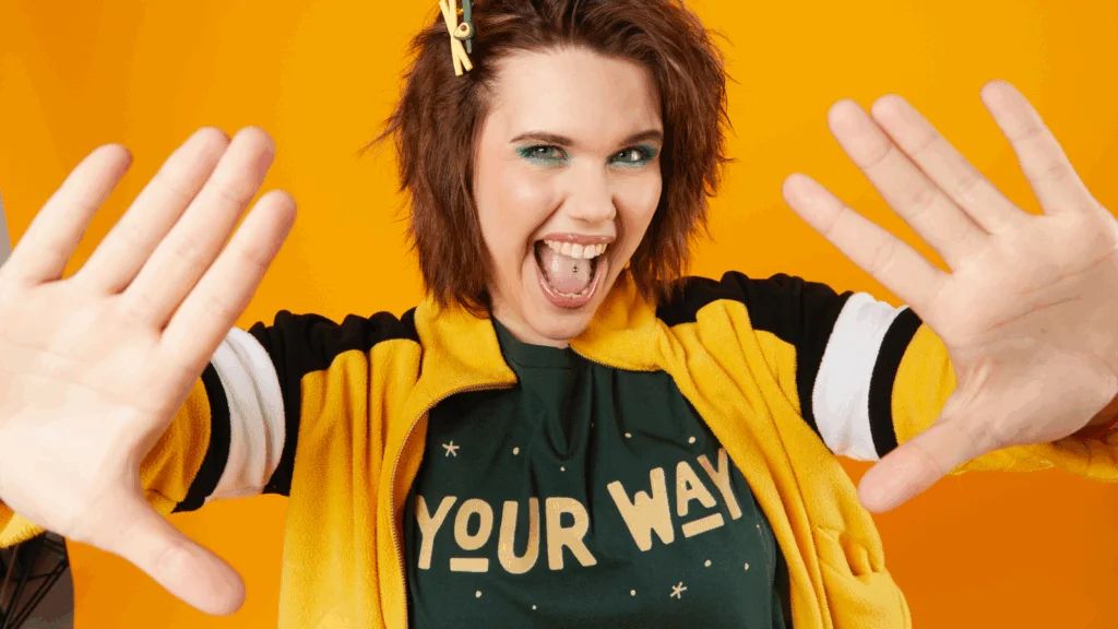 A woman with green eyeshadow smiling broadly, wearing a yellow and black jacket and a "Your Way" t-shirt.