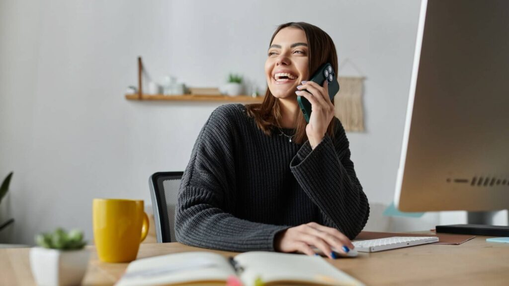 A smiling woman working on a desktop computer and making a phone call.