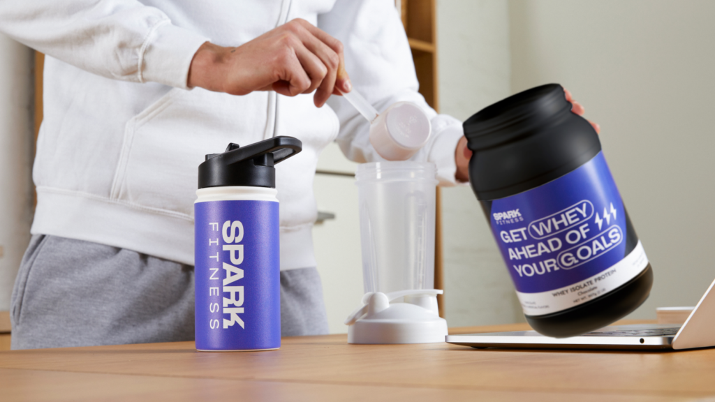 A person in a white hoodie prepares a protein shake at a table. A “SPARK FITNESS” bottle and whey protein container are visible.